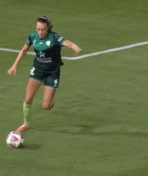 A Woman In A Green Jersey That Says University Of Canberra Kicks A Soccer Ball