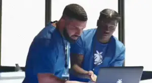 Two Men In Blue Shirts Are Looking At A Laptop .