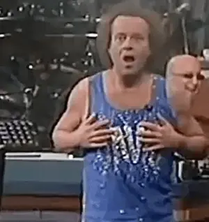 A Man In A Blue Tank Top Is Holding His Chest And Making A Face .