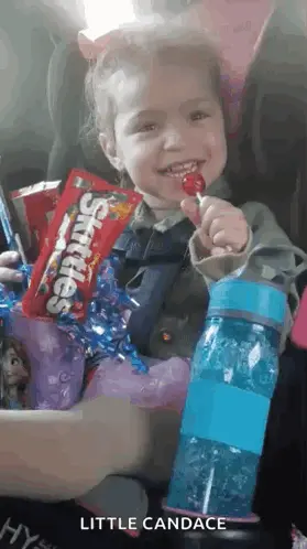 A Little Girl Is Sitting In A Car Seat Holding A Lollipop And A Bag Of Skittles