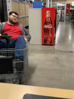 A Man Sits In A Shopping Cart In Front Of A Coca Cola Fridge
