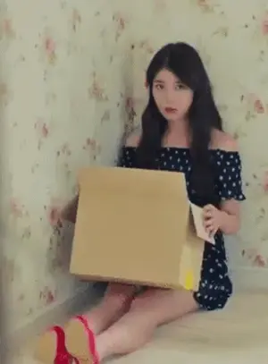 A Woman Is Sitting On The Floor Holding A Cardboard Box And A Piece Of Paper .