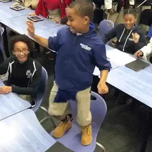 A Boy In A Blue Jacket Is Dancing In A Classroom While Other Children Sit At Tables .