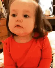 A Baby Girl In A Red Shirt Is Sitting On A Bed Making A Funny Face .