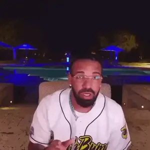 A Man With A Beard Wearing Glasses And A Baseball Jersey Is Sitting In Front Of A Pool At Night .