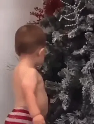 A Shirtless Baby Is Standing Next To A Christmas Tree .