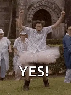 A Man In A Tutu Is Standing In The Grass With His Arms In The Air And A Group Of People Behind Him .