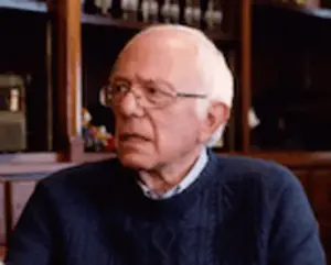 A Man Wearing Glasses And A Blue Sweater Is Sitting In Front Of A Bookcase .