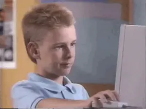 A Young Boy In A Blue Shirt Is Typing On A Laptop Computer .