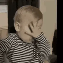 A Baby In A Striped Shirt Is Covering His Face With His Hands .