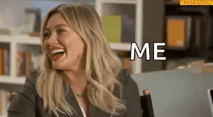 A Woman Is Laughing And Saying `` Me '' While Sitting In Front Of A Bookshelf .